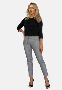 Czarna, półdopasowana bluzka z zebranym dekoltem; szare, w kratę, zwężane spodnie; modelka w czarnych, odkrytych sandałach na obcasie. Gładkie szare tło.