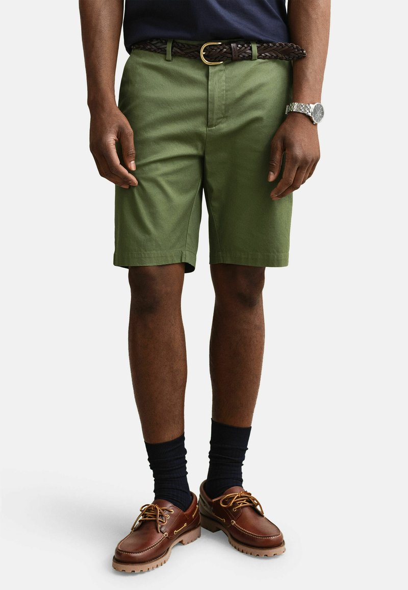 Hombre con pantalones cortos color verde oliva con un cinturón trenzado marrón, calcetines negros, zapatos náuticos de cuero marrón y un reloj de pulsera plateado, de pie.