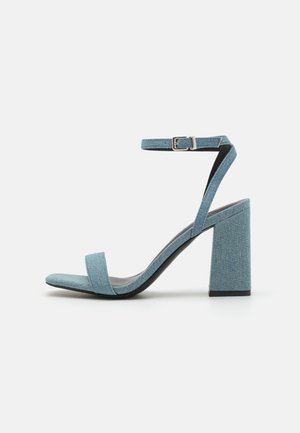 Sandale à talon haut en denim bleu à bout ouvert, avec un talon bloc, une bride de cheville et une bande à orteil simple et plate. Présente un tissu texturé et une semelle noire.