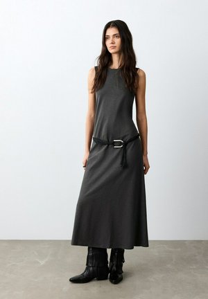 Frau mit langem dunklem Haar, die ein ärmelloses, dunkelgraues Maxikleid mit einem schwarzen Gürtel und schwarzen Stiefeln mit Fransen trägt, steht auf einem grauen Boden.