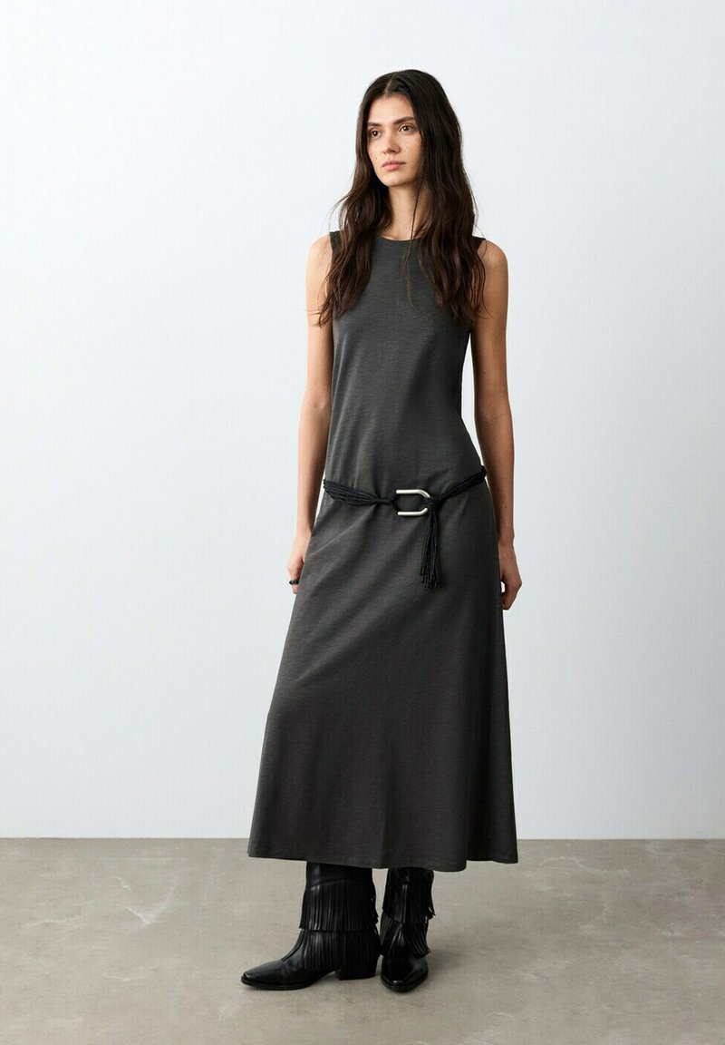 Femme aux longs cheveux foncés portant une robe maxi sans manches gris foncé avec une ceinture noire et des bottes à franges noires, debout sur un sol gris.