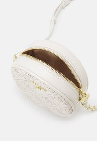 Kulatá kožená taška v krémové barvě s perforovaným vzorem, zlatým zipem a řetízkem jako zdobným akcentem. Otevřený zip odhaluje hnědou podšívku.