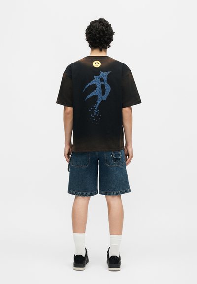 Άνδρας με την πλάτη γυρισμένη, φοράει μαύρο μπλουζάκι tie-dye με μπλε αφηρημένο σχέδιο και κίτρινο πρόσωπο με χαμόγελο, τζιν σορτς, λευκές κάλτσες και μαύρα αθλητικά παπούτσια.