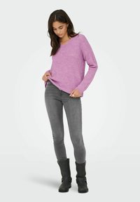 Femme portant un pull rose à col en V, un jean skinny gris, et des bottes noires mi-mollet, debout devant un fond clair uni.