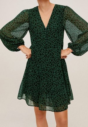 Frau trägt ein knielanges grünes Kleid mit schwarzem Leopardenmuster und transparenten langen Puffärmeln, Hände an den Hüften.