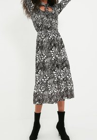 Robe à motifs noir et blanc avec un corsage smocké, des manches longues, un détail en découpe, et un ourlet à volants, portée avec des bottines noires à la cheville.