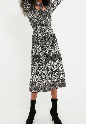 Robe à motifs noir et blanc avec un corsage smocké, des manches longues, un détail en découpe, et un ourlet à volants, portée avec des bottines noires à la cheville.