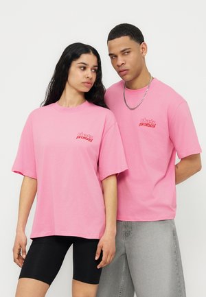 Two people wearing matching pink "chaos protocol" T-shirts; one wearing black shorts, the other gray jeans with a chain necklace.