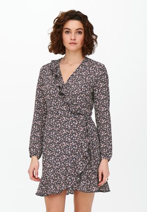 Femme aux cheveux bruns bouclés portant une robe portefeuille noire à manches longues avec un motif floral et des détails en volants sur fond blanc.
