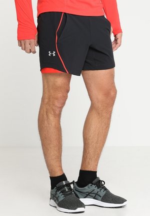 Benen van een persoon die zwarte en rode Under Armour shorts draagt, zwarte enkelsokken, grijze en zwarte hardloopschoenen en een rood topje met lange mouwen.