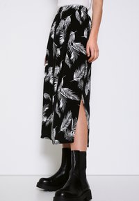 Nainen, joka on pukeutunut musta-valkoiseen lehtikuosiseksi midi-hameeseen, jossa on sivureikä, ja mustiin paksupohjaisiin nilkkasaappaisiin, seisoo valkoista taustaa vasten.