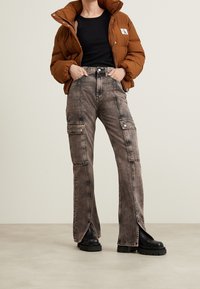 Chaqueta acolchada marrón con un cuello alto, top negro sin mangas y pantalones cargo grises con aberturas laterales y bolsillos, combinados con botas negras gruesas.