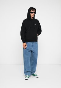 Sort bomulds-hættejakke med en forlommer og snørebånd, parret med lyseblå overdimensionerede denimjeans og grønne og hvide lavtoppede sneakers.