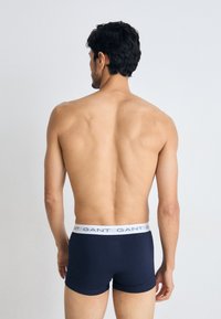 Marineblå boksershorts med en hvit elastisk midjebånd som har merkevarenavnet. Glatt tekstur, tettsittende design, bakside av modellen.