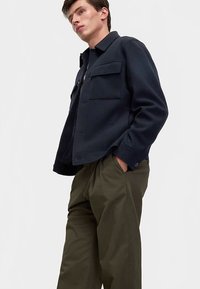 Granatowa kurtka z mieszanki wełny z zapinanym na guziki przodem, dwoma kieszeniami na piersi i kołnierzem. Połączona z luźnymi, oliwkowozielonymi spodniami.