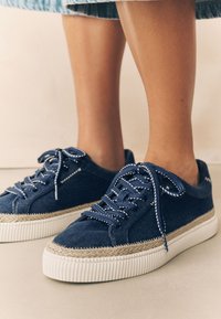 Marineblå lærreds-sneakers har en hvid tekstureret sål, dekorative syninger og fletdetaljer. Snørebåndene er blå med hvide accenter.