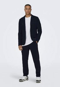 Donkere blazer over een wit t-shirt, gecombineerd met donkere broeken en sneakers. Glad materiaal, slim fit, met minimalistische ontwerpelementen.