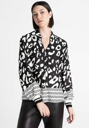Femme aux cheveux courts et foncés portant un chemisier noir et blanc à motifs avec des cœurs sur les poignets et un pantalon noir sur fond blanc.