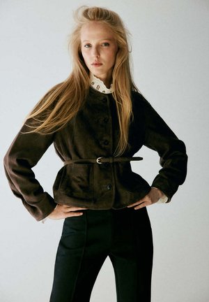 Mujer con cabello largo y rubio que lleva una chaqueta marrón oscuro con cinturón sobre una blusa blanca con cuello de encaje y pantalones negros, de pie con las manos en las caderas.