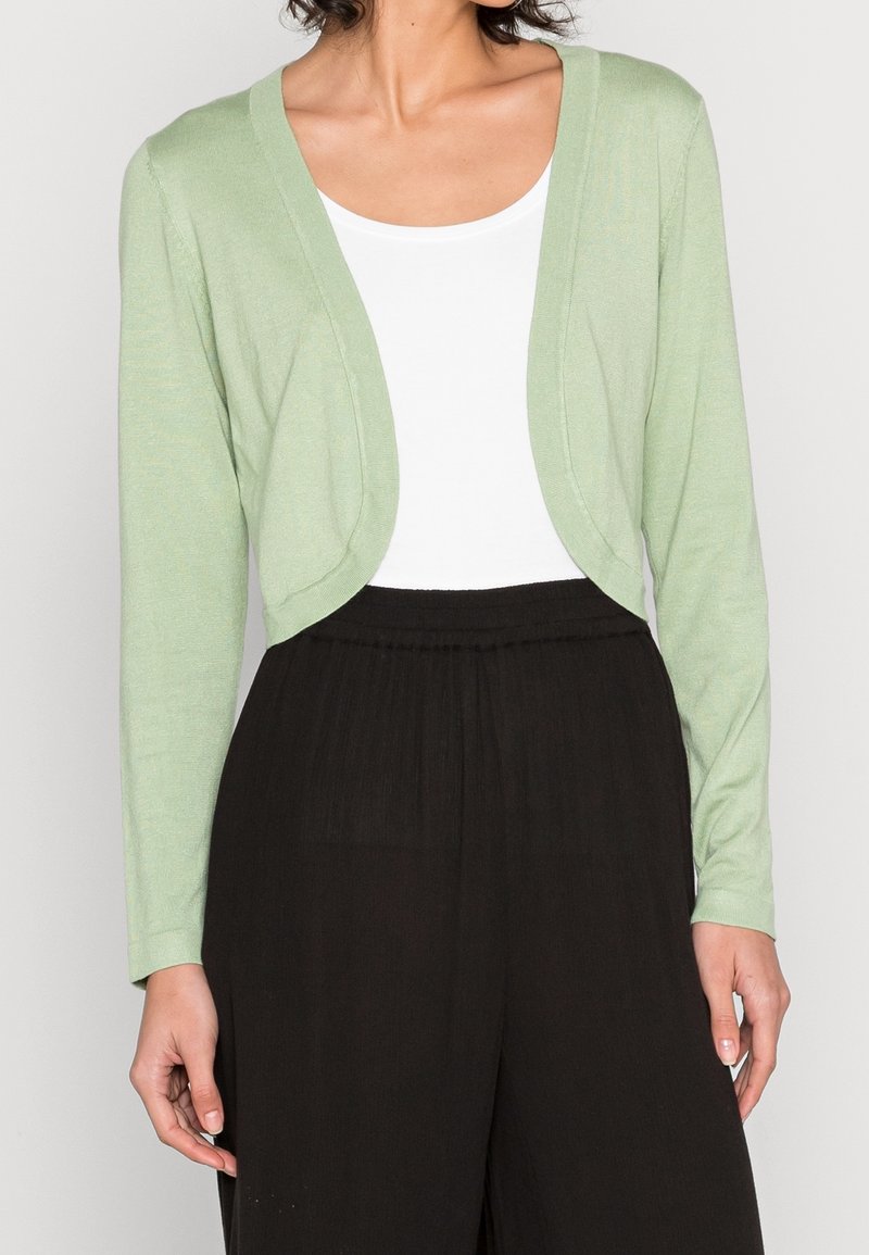 Vaaleanvihreä lyhyt neuletakki, jossa pitkät hihat, avoin etuosa ja pehmeä pinta, käytetty valkoisen topin ja mustien leveälahkeisten housujen päällä.