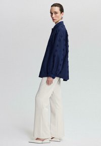Navy blue textured shirt with puffed sleeves, featuring a button closure, paired with wide white trousers and pointed white shoes.