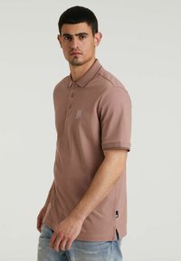 Polo tričko v prachovo ružovej farbe, vyrobené z bavlneného zmesi, s golierom, tromi gombíkmi, krátkymi rukávmi a logom na ľavej strane hrudníka.