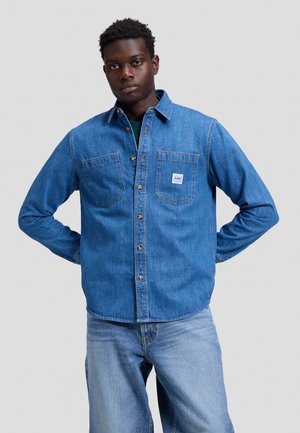 Joven con piel oscura y cabello rizado corto lleva una camisa azul de mezclilla con botones y vaqueros azul claro, de pie con las manos detrás de la espalda.