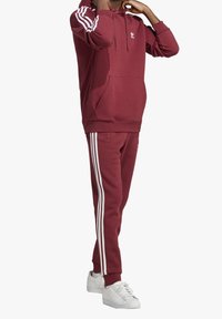 Bordeaux mikina s bílými pruhy na rukávech, klokání kapsa a odpovídající bordeaux kalhoty se bočním pruhem, doplněné bílými teniskami.