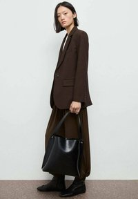 Blazer marrone su misura, pantaloni marroni morbidi, borsa a spalla nera in pelle con dettagli dorati e stivaletti neri alla caviglia con lacci, su uno sfondo neutro.