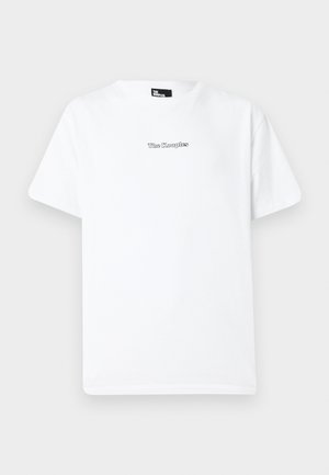 Giovane uomo in piedi, indossa una t-shirt bianca con stampa grafica, pantaloni neri a gamba larga con accessorio a catena e scarpe nere, su sfondo chiaro e uniforme.