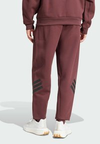 Pantalons de survêtement bordeaux à coupe décontractée, avec des accents noirs à trois rayures sur la jambe gauche, et une poche latérale pour plus de fonctionnalité.