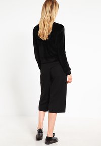 Svart fløyelsjakke med cropped design og lange ermer, parret med svarte culottbukser. Skoene har glansfullt svart materiale med runde tær.