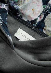 Tuvumā pelēka apģērba apkakle, kas parāda ziedu audumu iekšpusē un baltu Ted Baker zīmola etiķeti, kas iestrādāta apkaklē.