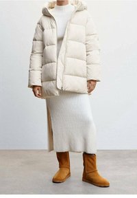 Béžová prošívaná bunda s kapucí, s prošívanými částmi a předním zipem. Nošena přes bílý žebrovaný svetr a hnědé kotníkové boty.
