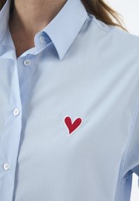 Lyseblå skjorte med korte ærmer og knaplukning, prydet med et rødt broderet hjerte på venstre side. Glat tekstur og klassisk kravedesign.