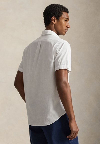 Short-sleeve white textured shirt with a collar, back pleat detail, and curved hem. Worn with navy blue pants, viewed from the side.