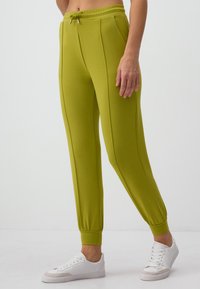 Grüne Jogginghosen aus weichem Stoff mit einem Kordelzugbund, schmal zulaufenden Säumen und zwei seitlichen Nähten; kombiniert mit weißen Turnschuhen.
