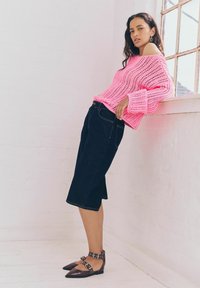 Maglione rosa a maglia aperta con maniche larghe, abbinato a culotte in denim scuro. Le scarpe sono ballerine con punta a punta e dettagli con fibbia in marrone scuro.