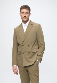 Hellbrauner doppelt gefütterter Anzug mit Reverskragen, zwei vorderen Taschen und passender Hose. Weißes Hemd darunter. Glatte Textur.