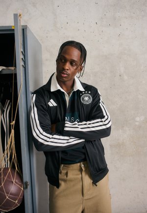 Schwarze Sportjacke mit weißen Streifen, kombiniert mit einem dunkelgrünen gestreiften Shirt. Beigefarbene Hose; im Hintergrund sind ein Spind und ein Basketball zu sehen.