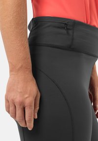 Zwarte leggings van een gladde, rekbare stof, met een hoge tailleband en een klein ritszakje aan de zijkant. De naden zijn zichtbaar.