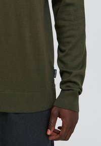 Heren olijfgroen gebreid sweater met ribbels aan de manchetten en onderkant, met een klein zwart label aan de zijkant. Zachte textuur en getailleerd ontwerp.