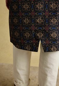 Melna rakstota auduma ar sarežģītiem zelta un zila dizaina elementiem. Līdz ceļgaliem veidota griezuma ar sānu šķēlumu, kopā ar krēma krāsas biksēm.