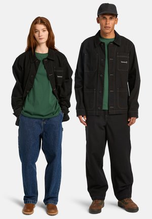 Zwei Modelle stehen nebeneinander und tragen schwarze Arbeitsjacken, grüne Hemden, weite Hosen und braune Schuhe; ein Modell trägt eine schwarze Mütze.