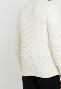 Maglione bianco a maglia strutturata con orlo e polsini a coste. Il design presenta un motivo a nido d'ape, evidenziando una vestibilità comoda e un tessuto morbido.