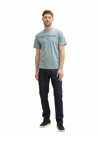 Helles blaues T-Shirt mit "TOM TAILOR"-Aufdruck; kombiniert mit dunklen Jeans und schwarzen Turnschuhen. Lässiger Schnitt, kurze Ärmel, minimalistisches Design.