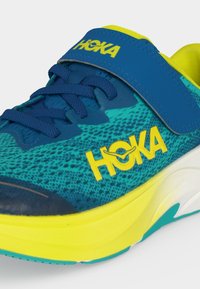 Μπλε και κίτρινο παπούτσι για τρέξιμο Hoka με υφασμένο επάνω μέρος, κορδόνια και αυτοκόλλητο λουράκι στο πάνω μέρος κοντά στον αστράγαλο.