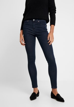 Femme portant un jean skinny bleu foncé, un pull noir à manches longues et des mocassins noirs, debout devant un fond clair uni.