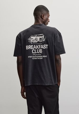 Mies mustassa t-paidassa ja housuissa, selkä meihin päin, paidassa on kahvilan luonnos ja teksti: "BREAKFAST CLUB, Aloita aamusi oikein voittaaksesi päivän."