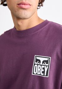 Lila pamut póló négyzet alakú fekete-fehér logóval, amelyen az "OBEY" szó és két stilizált szem látható. Egyszerű, kerek nyakkivágású.